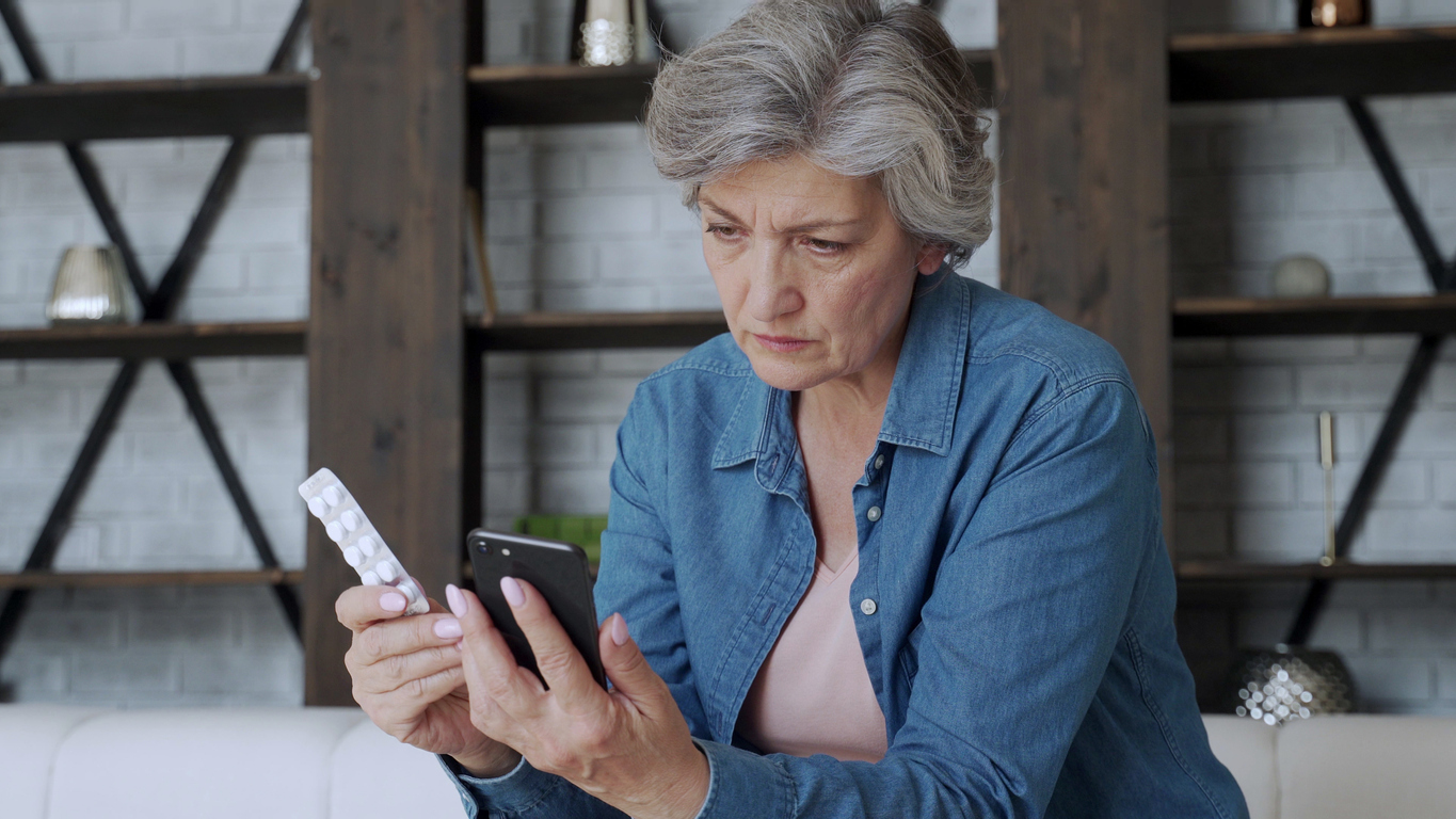 Elderly woman checks the recipe using her smartphone. Holds a smartphone in one hand and pill in the other